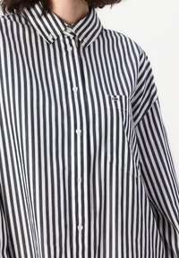 Camicia a righe con bottoni, a righe verticali blu navy e bianche, colletto e taschino sul petto con un piccolo dettaglio del logo.