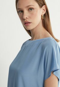 Ljusblå blus med kort ärm och rund halsringning, gjord av mjukt och flödande tyg. Slät textur med minimala designelement och rena linjer.