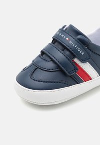 Námořnické modré tenisky z umělé kůže s bílou podrážkou, vybavené suchými zipy a červenobílými pruhy. Logo zobrazeno na pásku.