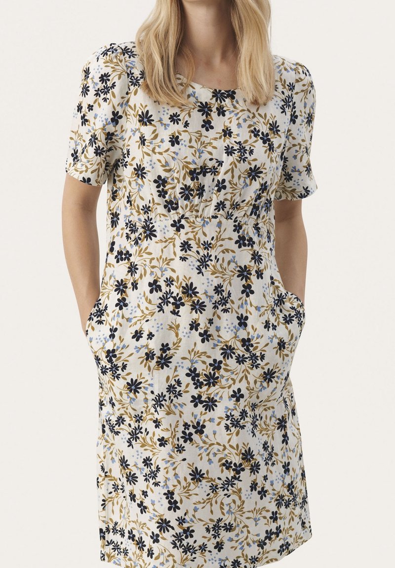 Femme portant une robe blanche courte aux manches courtes, longueur genou, avec des motifs floraux noirs et marron, mains dans les poches, debout devant un fond uni.
