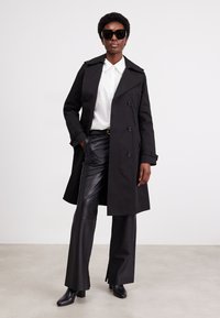 Schwarzer, doppelt-knöpfbarer Trenchcoat mit taillierter Silhouette, breiten Revers und Knopfdetails, kombiniert mit schwarzen Lederhosen und Stiefeln mit Absatz.