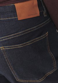 Tmavě modré džíny s hladkou denimovou strukturou, zdobené žlutým prošíváním a hnědou koženou štítkem s raženým nápisem "TNTUUM" na pásku.