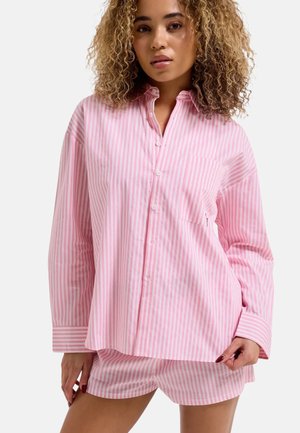 Woman with curly hair wearing a loose pink and white vertical striped button-up shirt and matching shorts against a plain background.