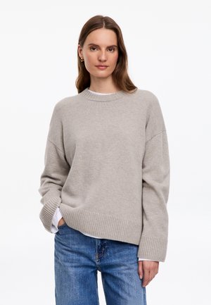 Femme aux longs cheveux bruns, portant des boucles d'oreilles créoles dorées, un pull oversize gris clair et un jean bleu, debout devant un fond blanc.