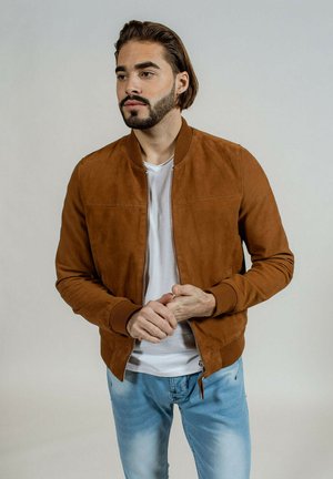 Veste bomber en daim marron avec une fermeture éclair, des poignets et un ourlet côtelés, portée sur un t-shirt blanc, associée à un jean bleu clair.