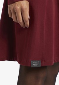 Abito bordeaux realizzato in tessuto morbido, con un'etichetta all'orlo che riporta i dettagli del marchio, e collant scuri a contrasto visibili.