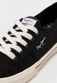 Czarny niski tenisówka z białymi sznurowadłami, białą gumową podeszwą oraz oznaczeniem "Pepe Jeans London" na języku i boku.