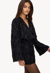 Zwarte jurk van textuurstof, diepe V-hals, lange klokmouwen, en gerimpelde taille. Bevat intrikate bloemmotieven door het hele ontwerp.