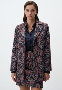 Blazer à motif floral dans des tons bleu foncé, rouge et gris, avec un col à revers classique et des manches longues, associé à une jupe coordonnée.