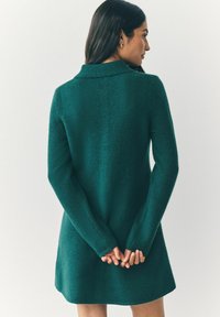 Zelené pletené šaty s golierom, dlhými rukávmi a voľným strihom. Textúrovaná látka. Modelka má ruky zložené za chrbtom.