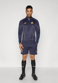 Marineblå sportsdress med hvite detaljer, glidelås foran, og Puma-logo. Inkluderer matchende shorts, knelange sokker, og fotballsko.
