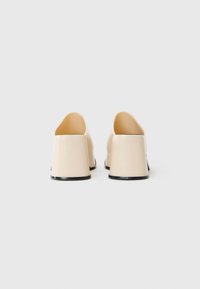Pantofi slip-on bej cu un finisaj neted, spate unghiular și talpă solidă neagră. Designul prezintă un spate deschis și un stil minimalist.