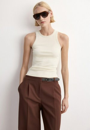 Blonde persoon met grote donkere zonnebril, mouwloos wit geribbeld topje, bruine broek met hoge taille en zwarte riem, staand tegen een witte muur.