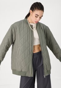 Prešívaná bomber bunda v olivovo zelenej farbe s rebrovanými manžetami, so zipsom vpredu a uvoľneným strihom, spárovaná s voľnými šedými nohavicami.