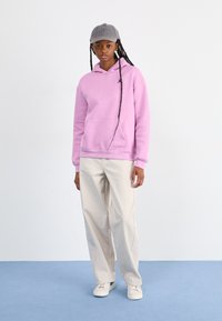 Lavender hoodie with a front pocket, paired with light beige wide-leg pants and white sneakers. Gray baseball cap completes the outfit.