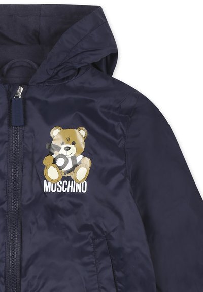 Giacca a vento blu navy con cappuccio, caratterizzata da un grafiche di un orso e il testo "MUSCHINO". Texture liscia e dettaglio della chiusura con zip visibile.