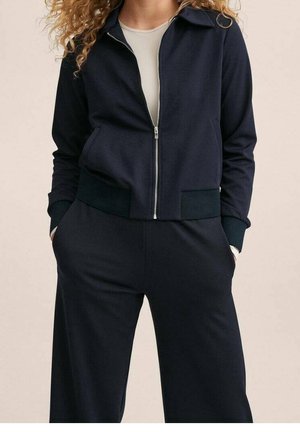 Femme aux cheveux bouclés portant une veste zippée bleu marine sur un haut beige et un pantalon bleu marine assorti, les mains dans les poches, debout devant un fond uni.