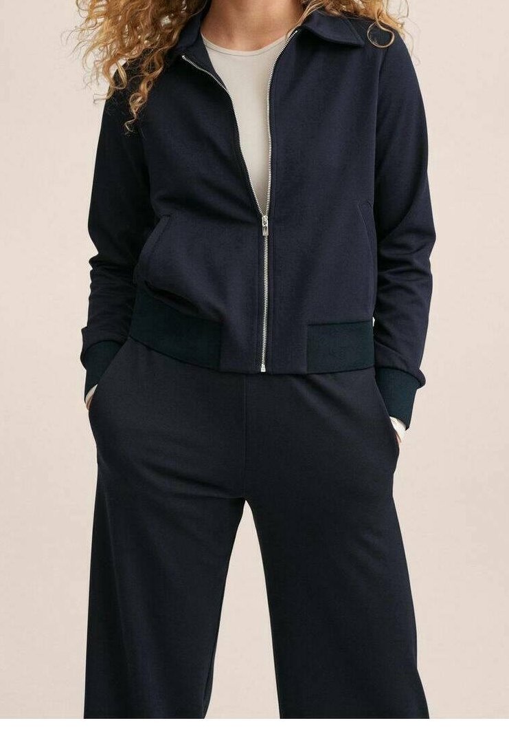Femme aux cheveux bouclés portant une veste zippée bleu marine sur un haut beige et un pantalon bleu marine assorti, les mains dans les poches, debout devant un fond uni.