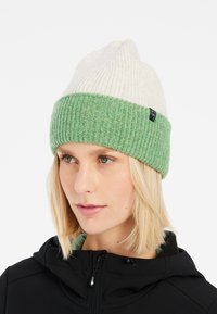 Knit beanie with a cream top half and a green ribbed lower band. Features a subtle brand tag on the side. Soft texture and fitted design.