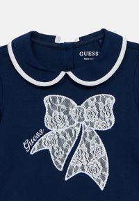 Ρούχο σε ναυτικό μπλε χρώμα με λευκό γιακά Peter Pan, διακοσμημένο με μία εντυπωσιακή λευκή δαντελωτή φιόγκο και το κεντημένο λογότυπο "Guess!".