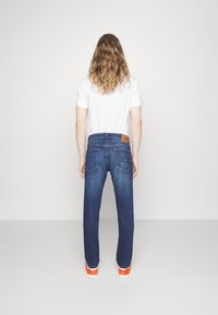 Mörkblå denimjeans med raka ben, en läderpatch baktill och kontrasterande sömmar. Kombinerade med en vit T-shirt.
