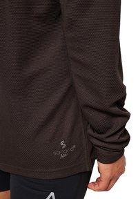 Mörkbrun träningstopp, strukturerad textil, med en logotyp vid fållen. Långa ärmar med samlade manschetter, designad för komfort och rörlighet.