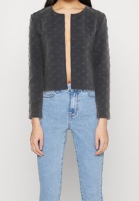 Chaqueta recortada de frente abierto en tejido gris oscuro texturizado con mangas largas. Combinada con jeans de color azul claro. Diseñada con un escote redondo.