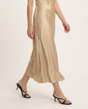 Kobieta idąca w beżowej satynowej sukience midi i czarnych sandałach na obcasie z odkrytymi palcami i złotą klamrą.