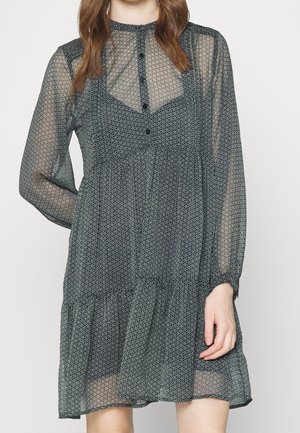 Frau trägt ein knieumspielendes, langärmliges, schwarzes, transparentes Kleid mit kleinen weißen Sternmustern und knöpfbarem Ausschnitt, posiert mit einer Hand an der Seite.