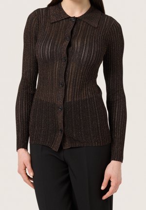 Schwarze gerippte Strickjacke mit Kragen, die feine metallische Fäden enthält, Knopfverschluss und lange Ärmel. Getragen über schwarzen Hosen.