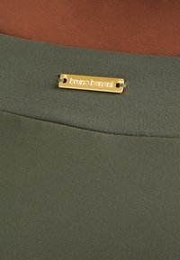 Olivgrüner Stoff mit glatter Textur, versehen mit einer goldenen Metallmarke, auf der "bruno banani" eingraviert ist.