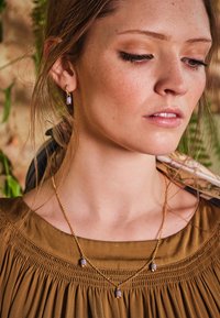 Guldhalsband med tre lila pärlor och matchande örhängen. Klänningen är brun med en samlad textur som kompletterar smyckena.