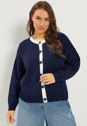 Donna che indossa un cardigan blu navy con bottoni e rifiniture bianche e jeans azzurri, posando davanti a uno sfondo chiaro semplice.