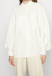 Camicia bianca oversize con colletto, maniche lunghe a palloncino, due tasche sul petto e una superficie liscia, abbinata a pantaloni beige chiaro.