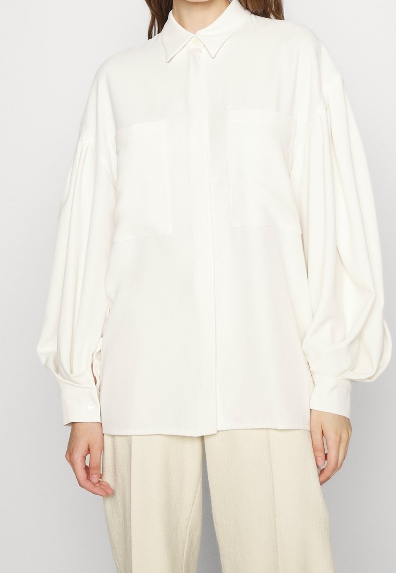 Camicia bianca oversize con colletto, maniche lunghe a palloncino, due tasche sul petto e una superficie liscia, abbinata a pantaloni beige chiaro.