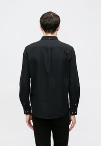 Cămașă neagră cu mânecă lungă și guler cu nasturi, având o pliură centrală în spate și detalii cu nasturi aurii pe manșete. Textură netedă.