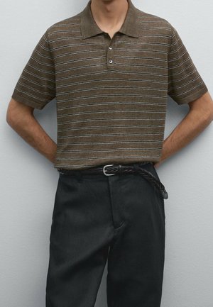 Mężczyzna ubrany w brązową koszulkę polo z krótkim rękawem w paski, do czarnych spodni z wysokim stanem i plecionym czarnym paskiem, stoi przy szarej ścianie.