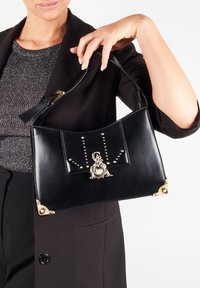 Mustan nahka käsilaukku, jossa on kaareva muotoilu, koristeltu kultaisilla metalliosilla ja niiteillä. Keskellä on koristeelline solki.