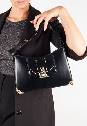 Borsa a tracolla in pelle nera con un design curvo, arricchita da accessori in oro e borchie. Presenta una chiusura decorativa al centro.