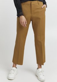 Pantalons marron ajustés à coupe droite, avec une taille plate et des poches latérales, associés à des baskets blanches sur un fond clair.