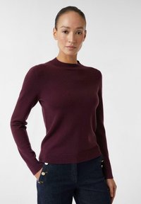 Vrouw met haar in een staart, die een getailleerde, donkerpaarse sweater met lange mouwen en een donkerblauwe broek met gouden knopen draagt, staand met één hand in de zak.