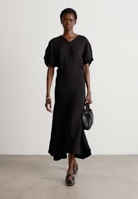Zwarte midi-jurk met korte pofmouwen, V-hals en een vloeiende rok. Model houdt een getextureerde zwarte handtas vast en draagt zwarte platte schoenen.