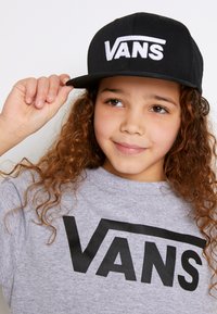 Cappellino snapback nero con il logo "VANS" ricamato in bianco, indossato da un bambino con capelli ricci, abbinato a una t-shirt grigia con un logo nero.
