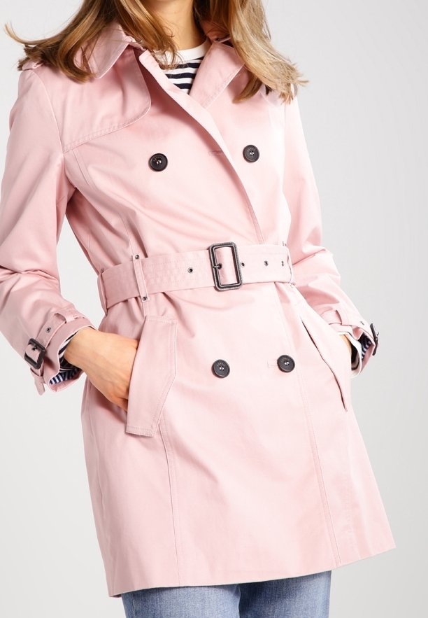 Ljust rosa trenchcoat med midjebälte, dubbelknäppning, stor krage och sidofickor. Tyget har en slät textur.