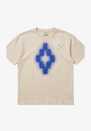 Maglietta beige a maniche corte con una forma a diamante geometrica blu pixelata stampata al centro e un piccolo tag bianco attaccato vicino al colletto.