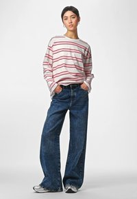 Woman standing with hands in pockets, wearing a white and red striped long-sleeve shirt, wide-leg blue jeans, and silver sneakers.