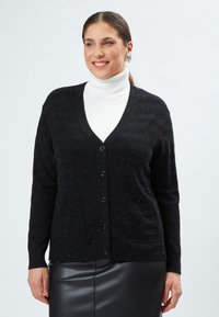 Cardigan nero con scollo a V, chiusura a bottoni, due tasche frontali e un knit testurizzato con sottili punteggiature. Indossato sopra un dolcevita bianco.