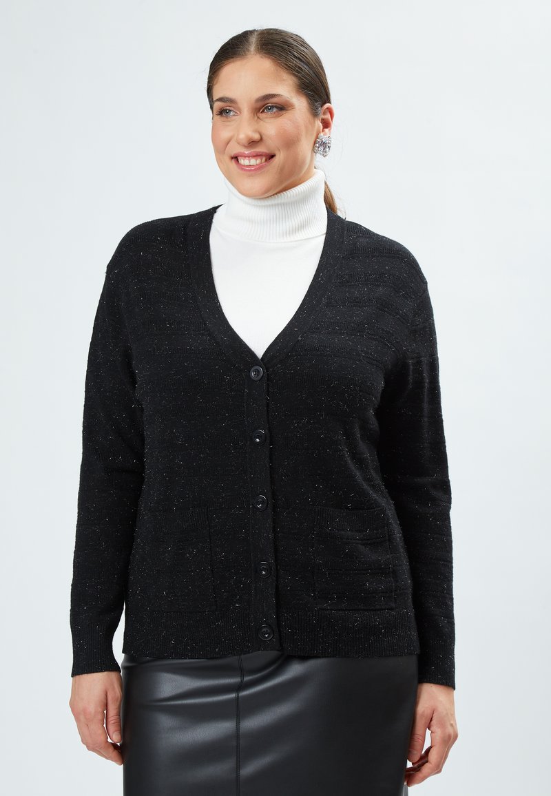 Cardigan nero con scollo a V, chiusura a bottoni, due tasche frontali e un knit testurizzato con sottili punteggiature. Indossato sopra un dolcevita bianco.