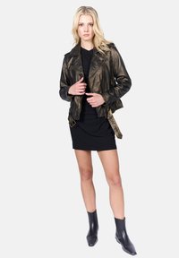Veste de biker en cuir noir avec une finition métallique, dotée de poches zippées, d'un col en pointe et d'accents à boucle. Portée sur une robe noire et des bottes.