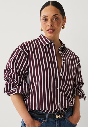 Striped shirt in dark burgundy and white, featuring a button-up front, collar, and rolled sleeves. Paired with high-waisted denim and a belt.